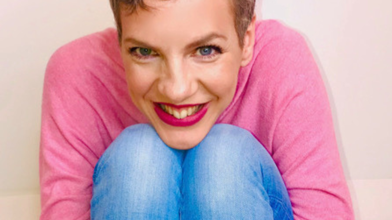 Francesca wears a pink jumper and jeans and smiles into the camera as she hugs her knees. Francesa has short brown hair and is wearing bright pink lipstick.