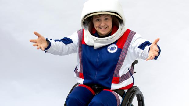 Jess Thom – a white woman with curly brown hair in front of a plain white background. Jess is smiling into the camera and her arms are outstretched. She is sitting in her wheelchair dressed as an astronaut in blue, white and red with a large white helmet on.