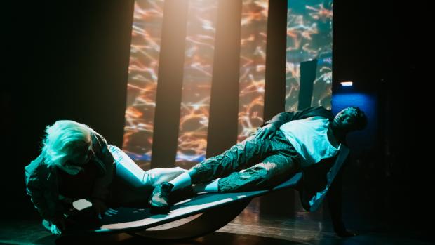 Two dancers are laying down on a rocking board, looking at each other. The board is tilted diagonally. The dancers are lit with a green light, contrasting with the background, where sound reactive visuals are projected on three screens and reflected on a mirror.