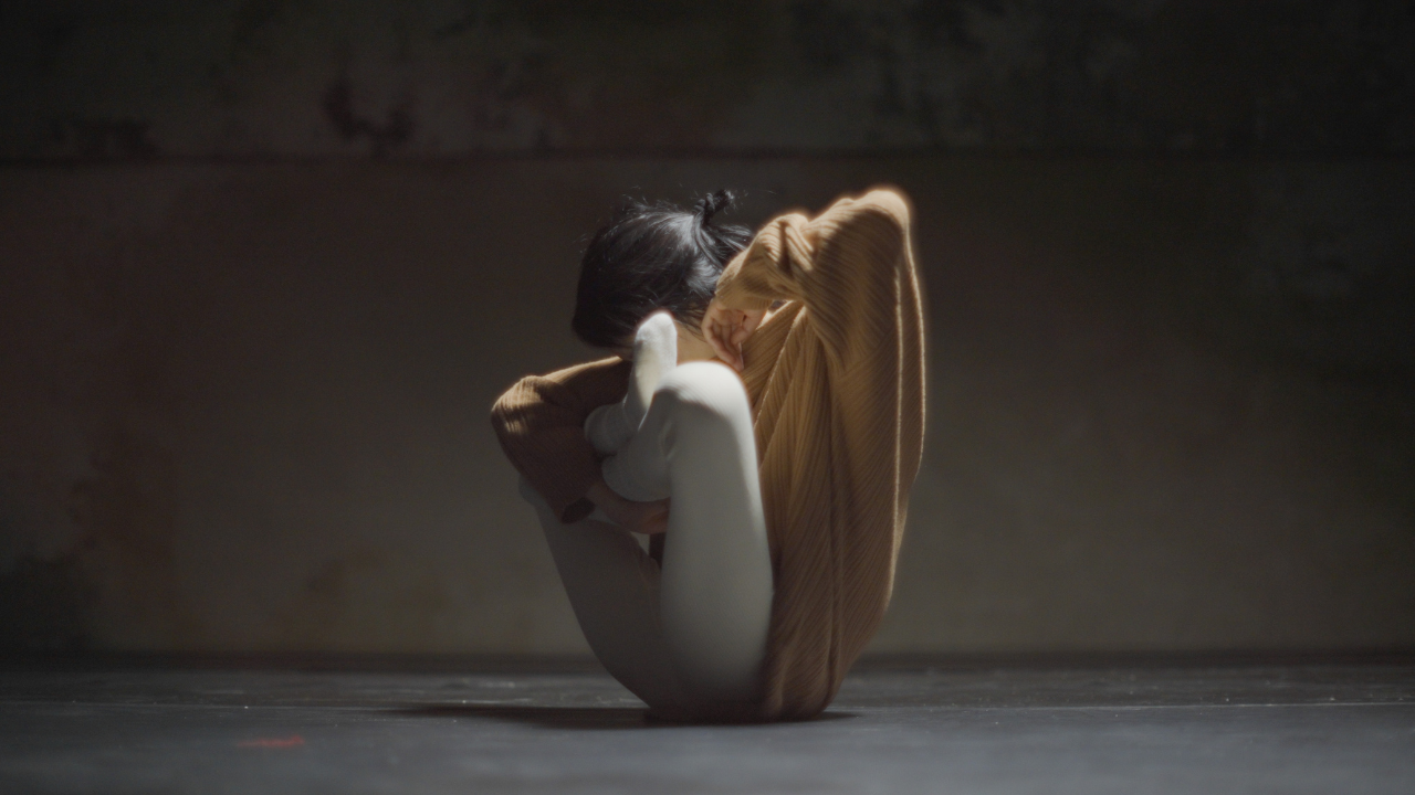 woman with legs crossed and held up to chest, head tucked in so we do not see her face as she balances on her bottom making an inverted triangular form in a dark room