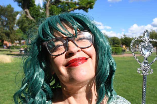 A woman with green curly hair in a glittery dress with a wand stands in a park smiling.