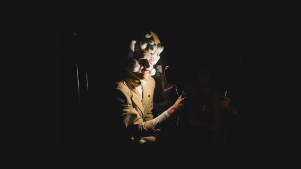 Meg Hodgson - MOONFACE, a drag creature with a Moon for a head stands smiling eerily at the camera. They are wearing a beige suit and white turtleneck, and are carrying a black desk lamp pointing at their face, and an extension cord.