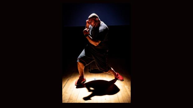 Rinkoo is standing on a wooden floor in a dark theatre space. He is a brown man with a bald head and black goatee beard. He wears a black t-shirt and shorts and bright red trainers. He is standing in profile, with his legs spread and hands up in a defensive boxing pose