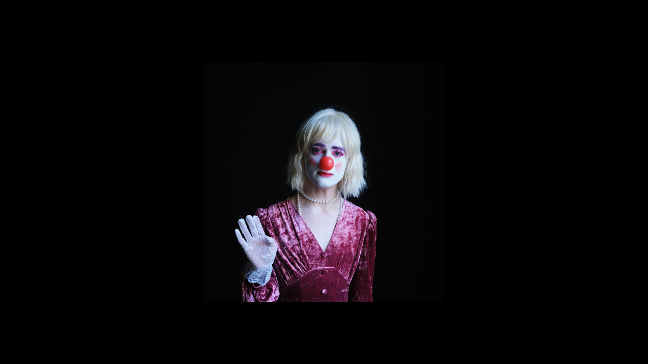 Jack Boal as his drag alter ego, Grace Fool. She has short blonde hair, a bright white face and a red clown nose. She is waving with her right hand and has a small smile. She is wearing a pearl necklace and purple dress.