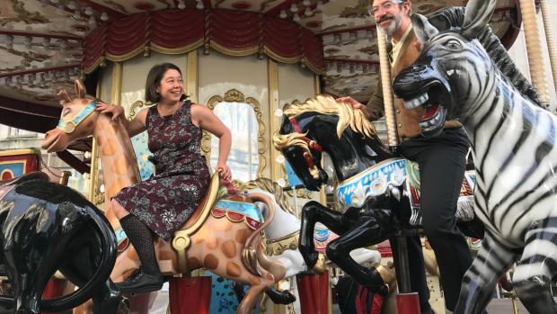 A man and a woman on a brightly coloured, old fashioned fairground carousel. The woman, Jo, sits on a camel wearing a maroon patterned dress and black knee length socks. The man, George, stands next to a black horse with a very excited zebra in front of him.