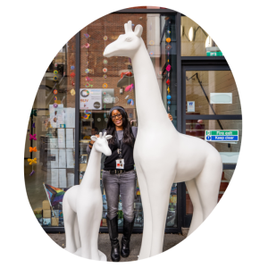 A giant and baby giraffe with a lady standing between them outside a shop front window