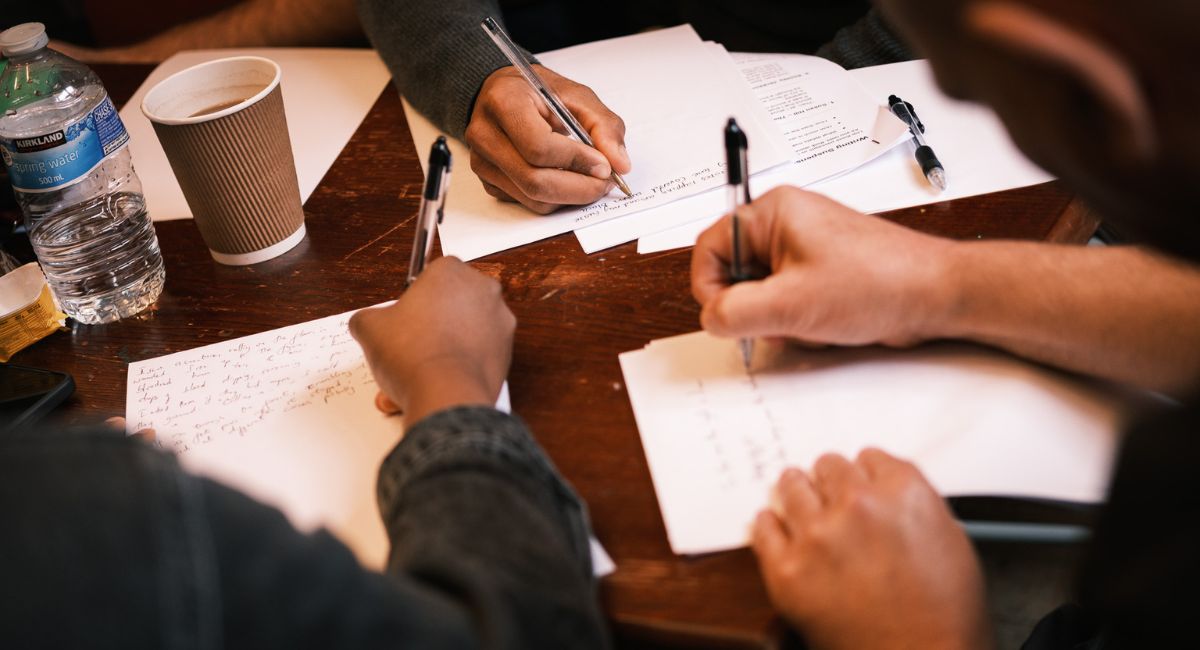 Three hands with different skin tones writing on blank paper, a coffee cup on the table