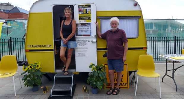 A woman and a man stand at the entrance of a bright yellow and white caravan labeled 