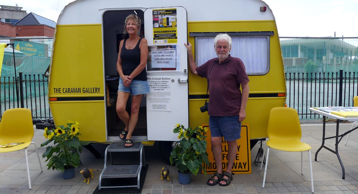 A woman and a man stand at the entrance of a bright yellow and white caravan labeled "The Caravan Gallery." The woman stands in the doorway wearing a black tank top and denim shorts, smiling at the camera, with a set of step stairs leading up to the door. The man stands beside the caravan in a maroon polo shirt and blue shorts, with a camera hanging around his neck.